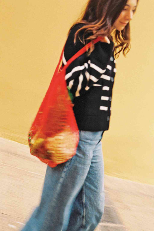 Woman walking past a yellow wall carrying a red mesh market bag filled with produce