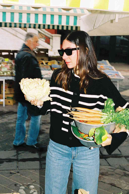 Woman in a striped cardigan at an outdoor market holding lettuce and a colander of fresh vegetables